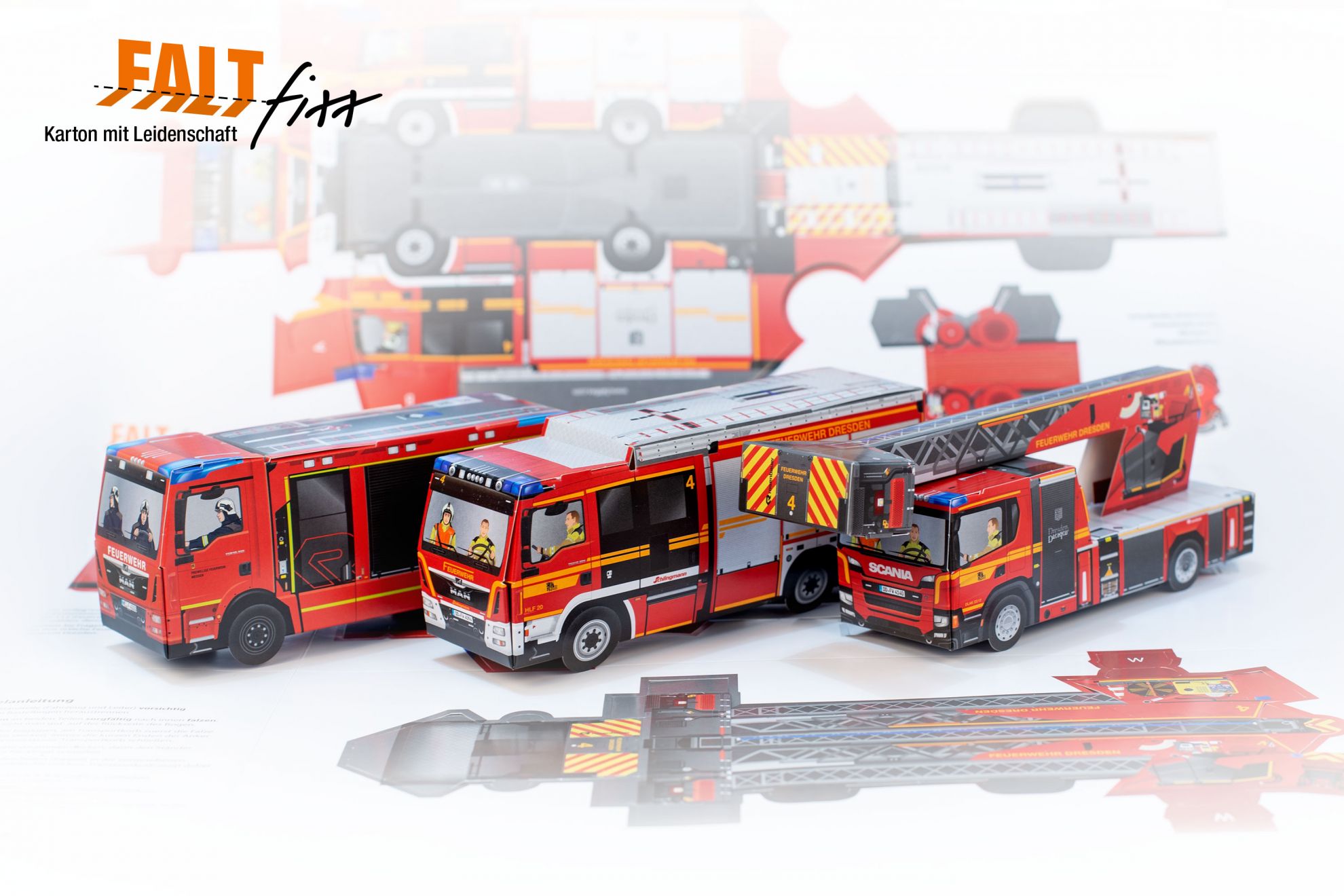 Drei zusammengesteckte Feuerwehrfahrzeuge aus Faltfixx Bastelbögen, im Hintergrund befindet sich ein ausgeklappter Bastelbogen